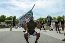Marine in green physical training uniform performs a front squat with barbell and plates in front of the Marine Corps Musuem with multiple uniformed Marines in the background.