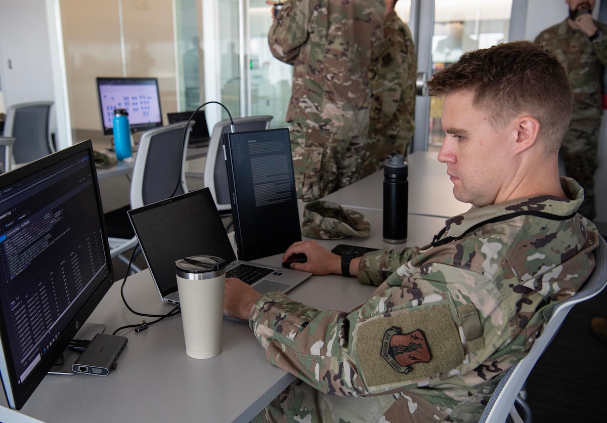 An airman in green uniform works at a computer desk