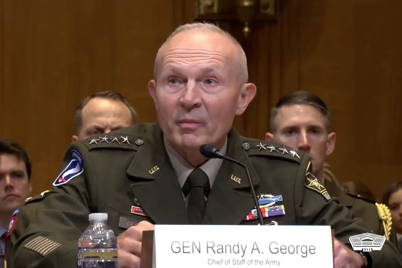 A man wearing a dress military uniform sits at a desk and speaks into a microphone. A sign in front of him reads "GEN Randy A. George." People in business attire sit behind him.