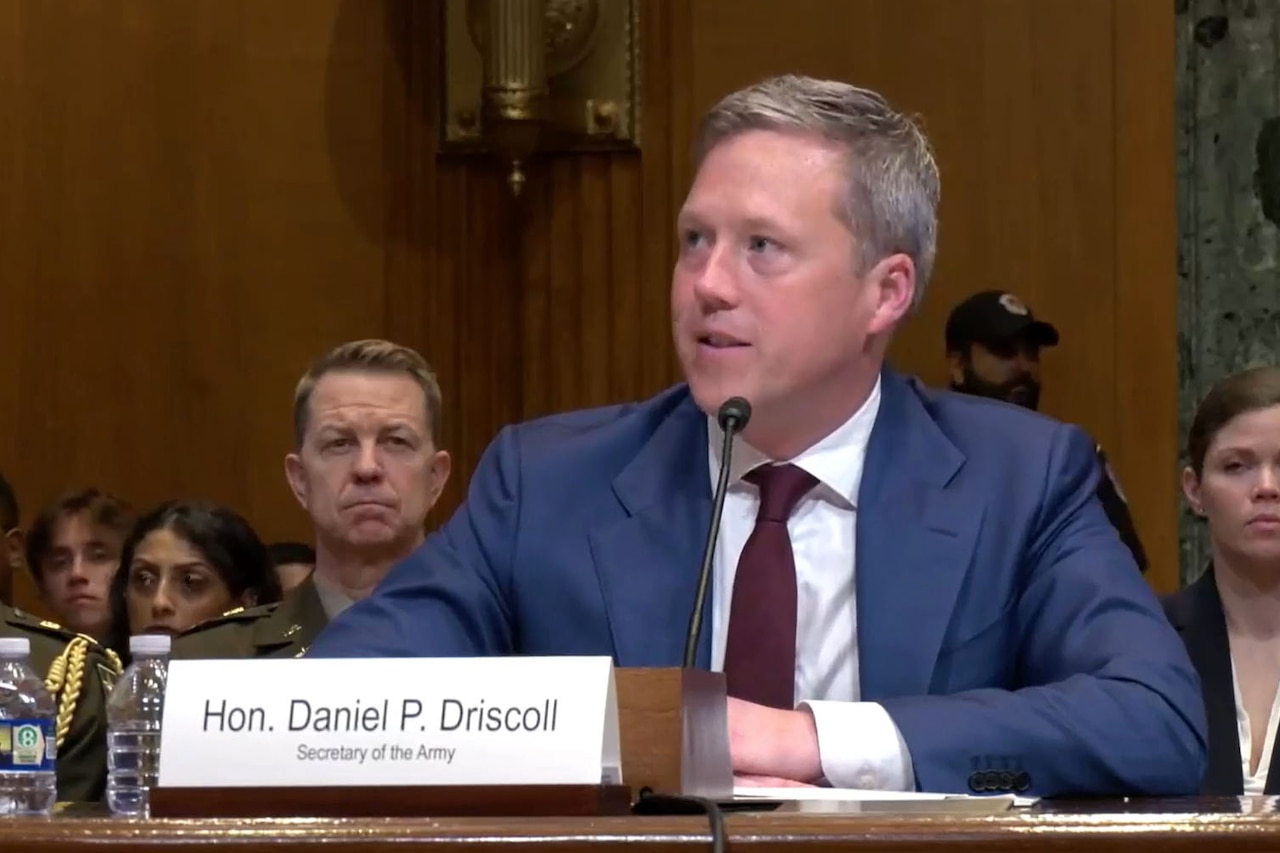 A man wearing business attire sits at a desk and speaks into a microphone. A sign in front of him reads, "Hon. Daniel P. Driscoll." More people in business attire sit behind him.