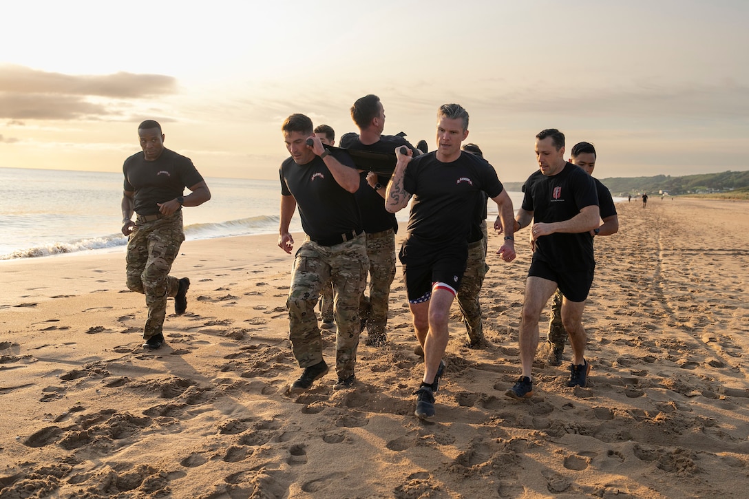 Defense Secretary Pete Hegseth helps carry a soldier on a litter while running with a group of troops on a beach.