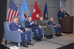 Men in chairs on stage as part of posture and readiness panel with another man at podium.