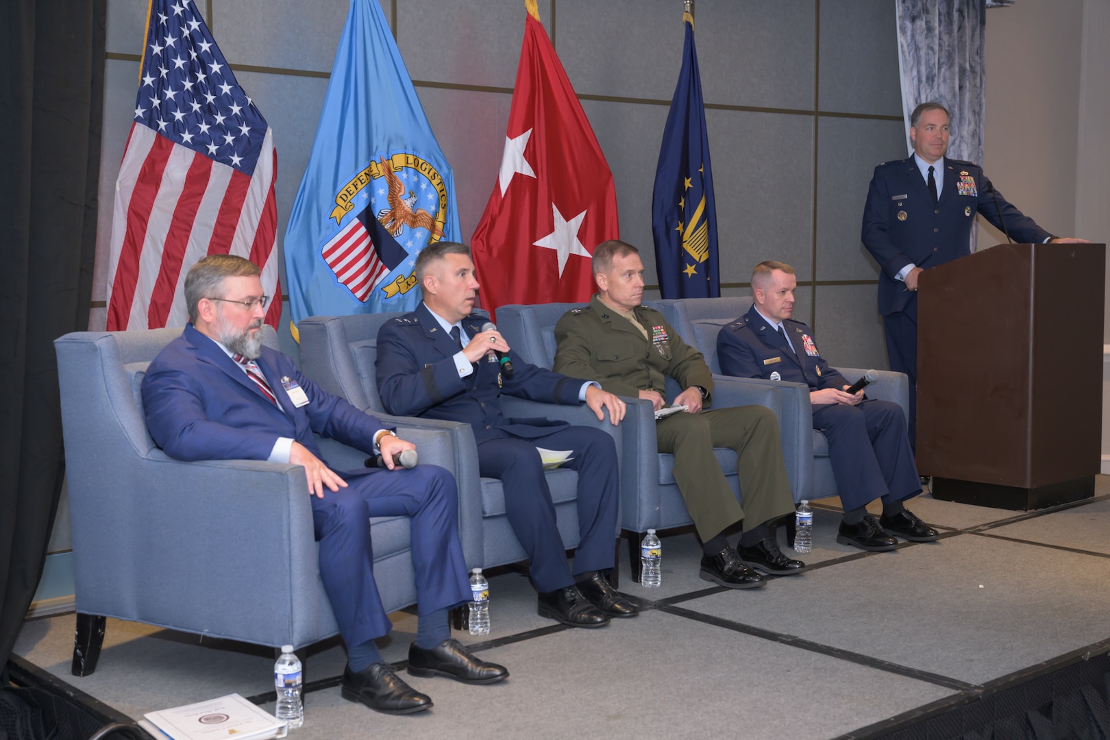 Men in chairs on stage as part of posture and readiness panel with another man at podium.