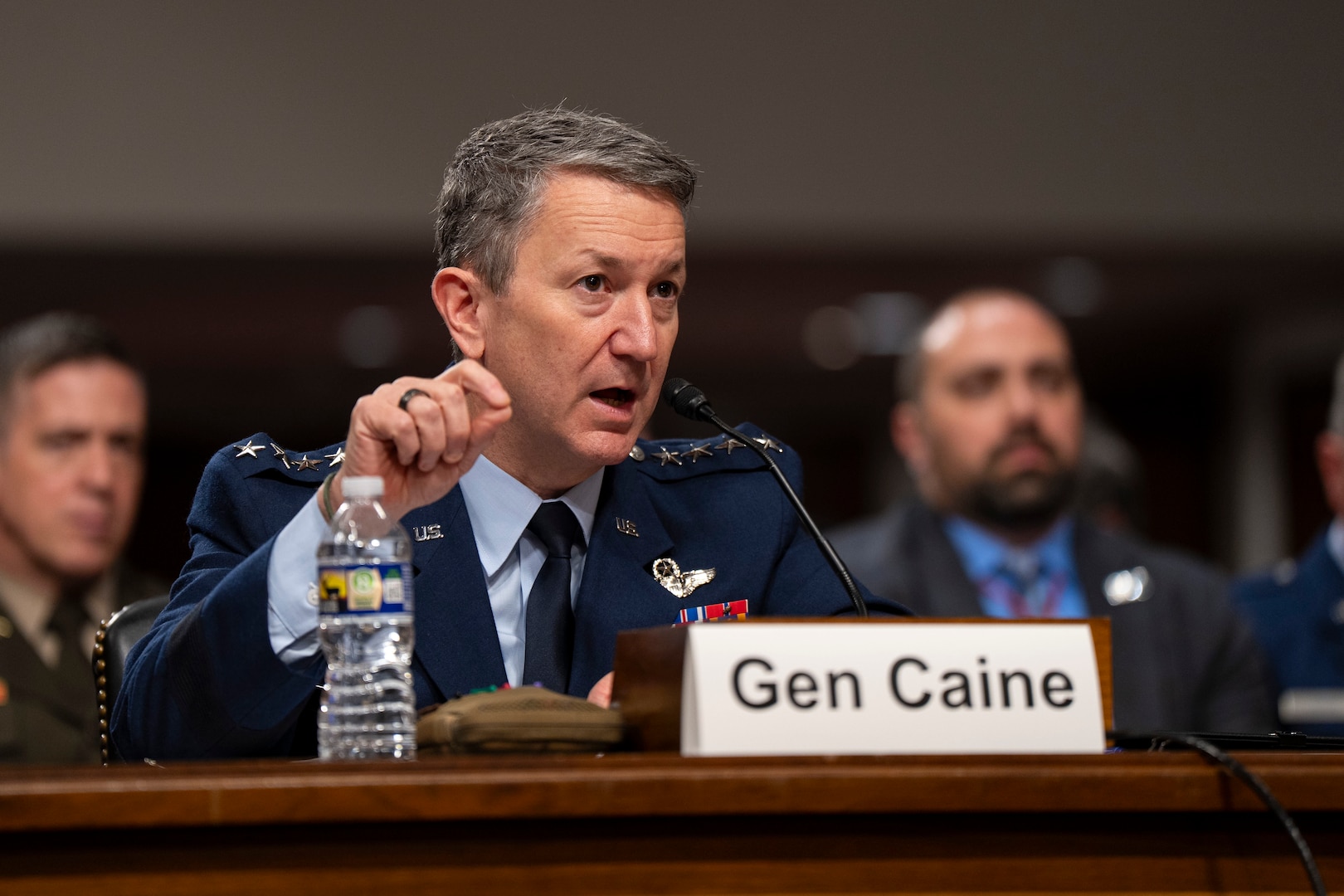 A man in a military suit speaks from behind a microphone while seated at a desk.
