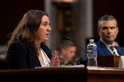 A woman speaks into a microphone while seated at a table in a room.