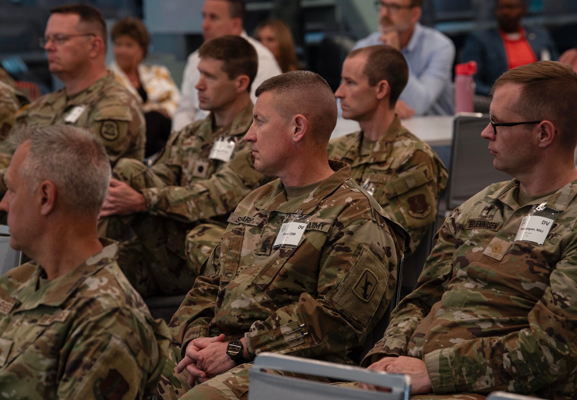 A group of military officials in green uniforms sit in the audience listening to a briefing.