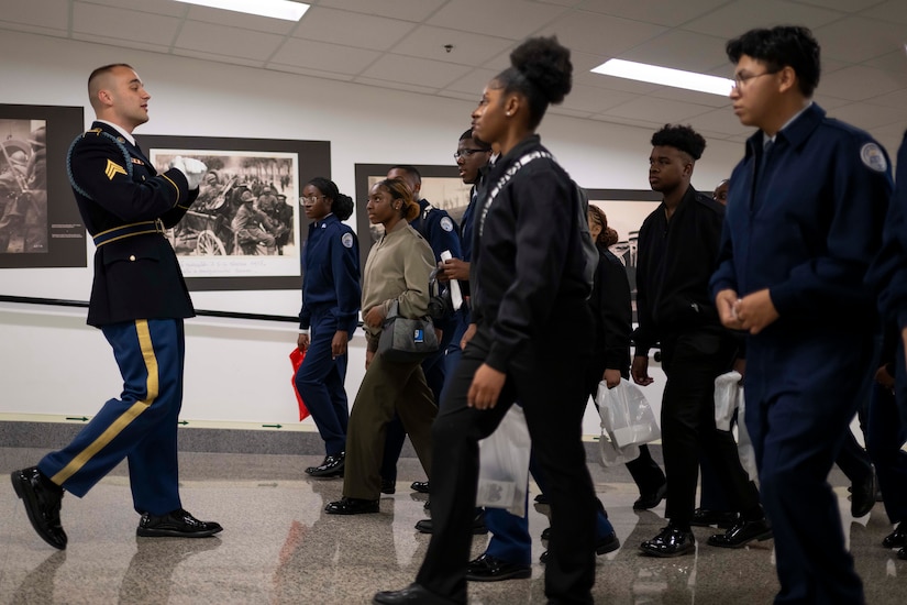 A soldier walks down a hallway backwards while speaking to youth walking forwards.