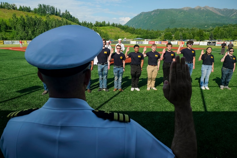 A sailor raises their right hand and faces a group of people wearing Navy T-shirts and doing the same on a field.