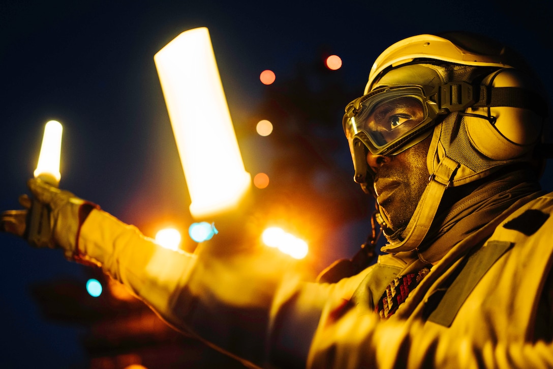 A close-up of a sailor wearing yellow protective gear while waving two neon lit batons.