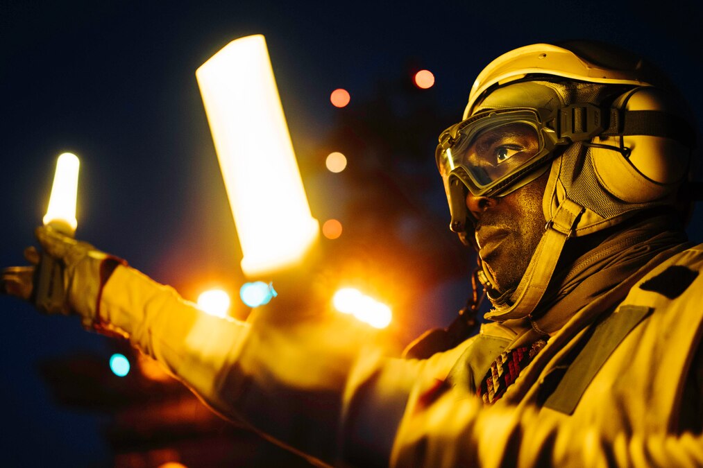 A close-up of a sailor wearing yellow protective gear while waving two neon lit batons.
