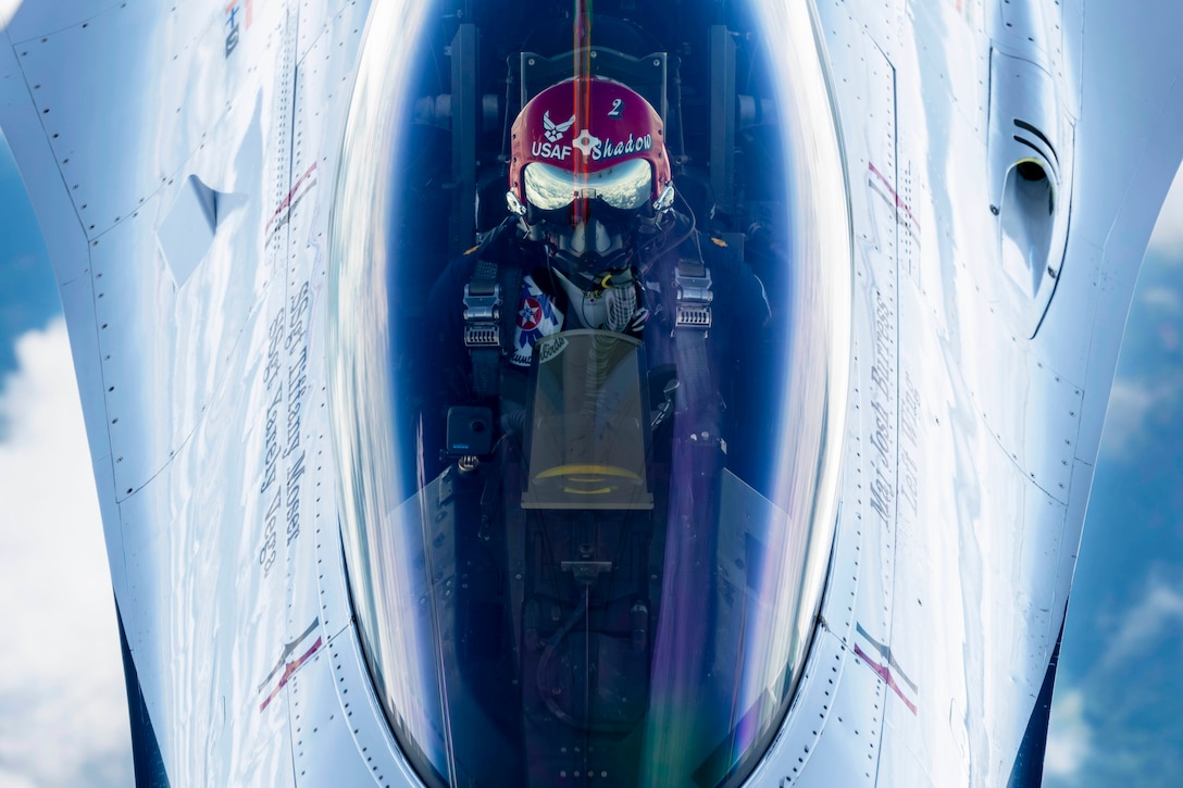 A close-up of an airman wearing protective gear flying downward in a cockpit against a cloudy, blue sky.