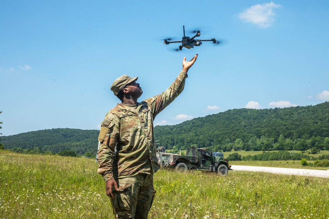 A soldier reaches for an unmanned aerial system while standing on a field under a blue sky.
