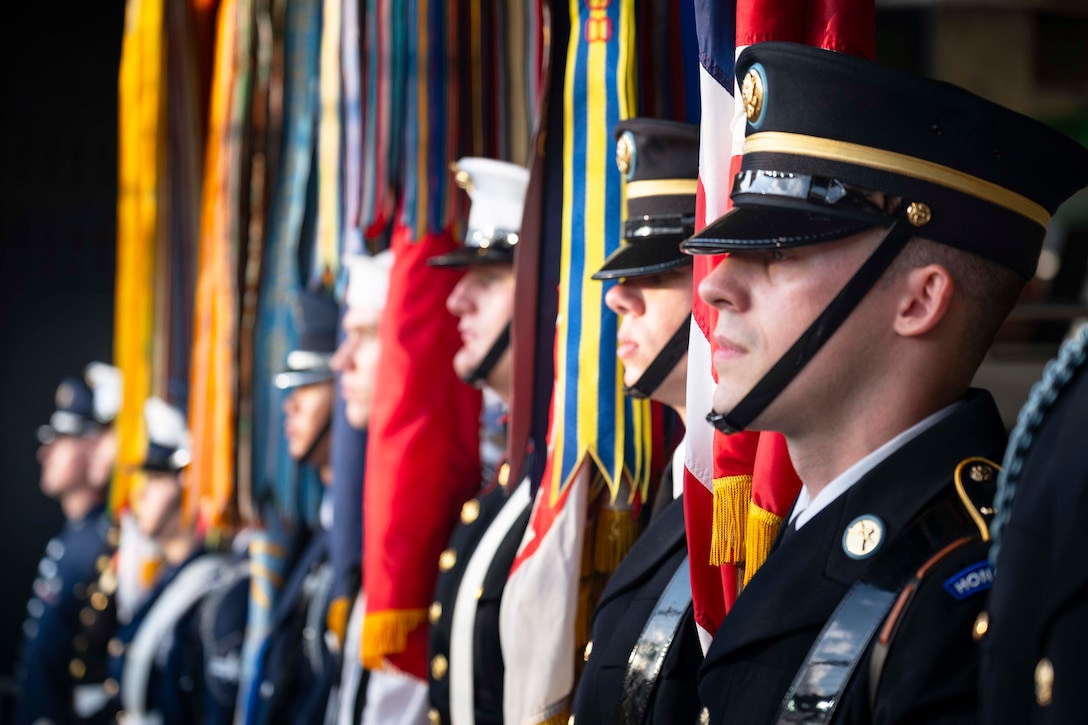 A side view of service members in ceremonial dress carrying flags while standing in formation.