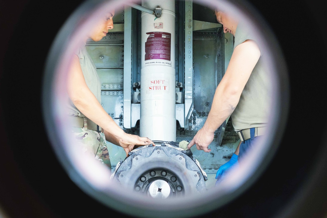 Two airmen face each other while working on a tire as seen through a circular window.