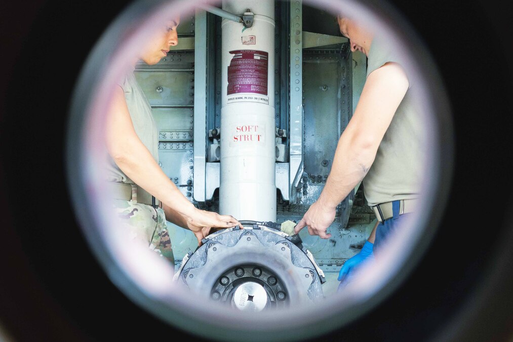 Two airmen face each other while working on a tire as seen through a circular window.