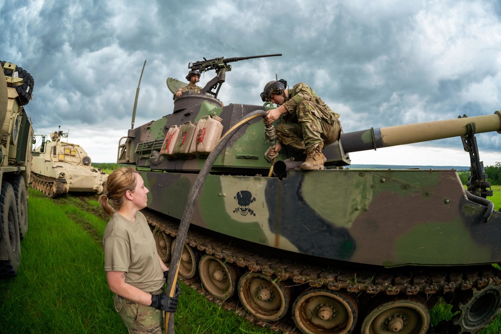 Two soldiers hold a hose while filling up a tank with gas in a field under a cloudy, gray sky.