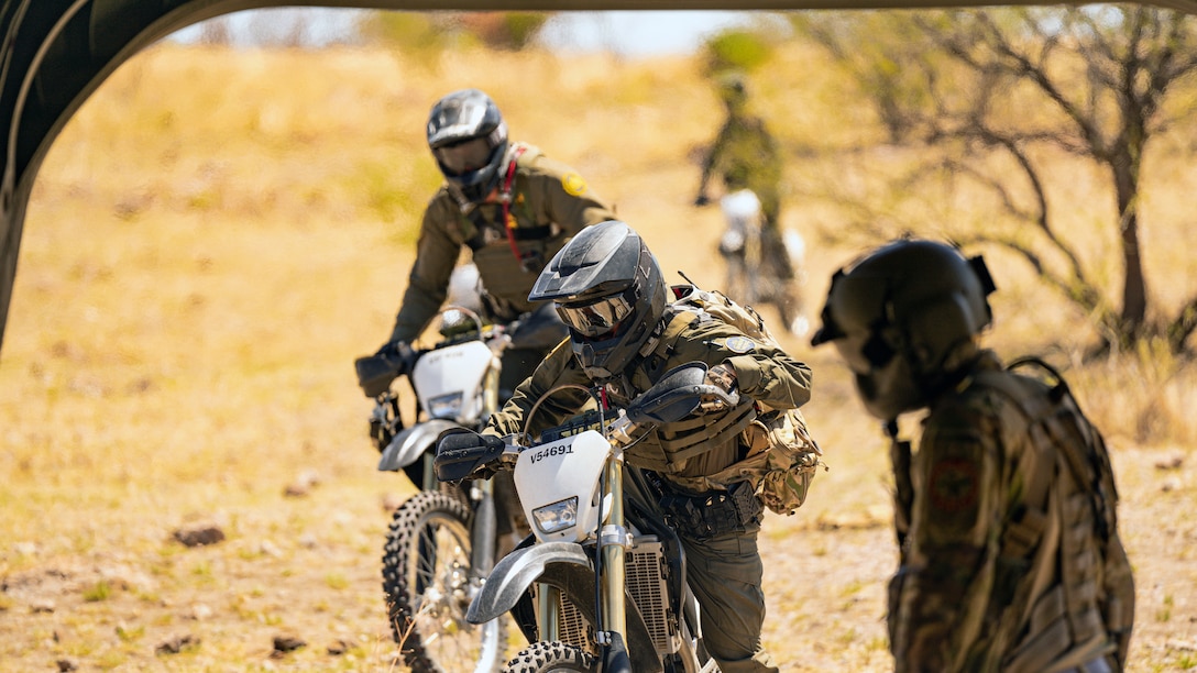 Soldiers and U.S. Customs and Border Protection personnel wearing helmets and goggles ride dirt bikes over remote terrain on a sunny day.