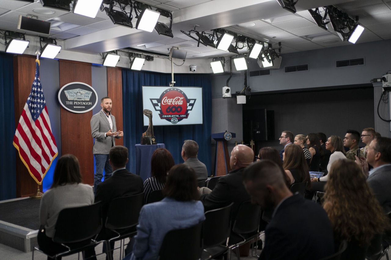 A man wearing business attire stands at the front of a room near a trophy. Behind him is an American flag, a sign that reads, "the Pentagon" and another sign that reads, "Coca-Cola 600."  In front of him are more than a dozen seated individuals.