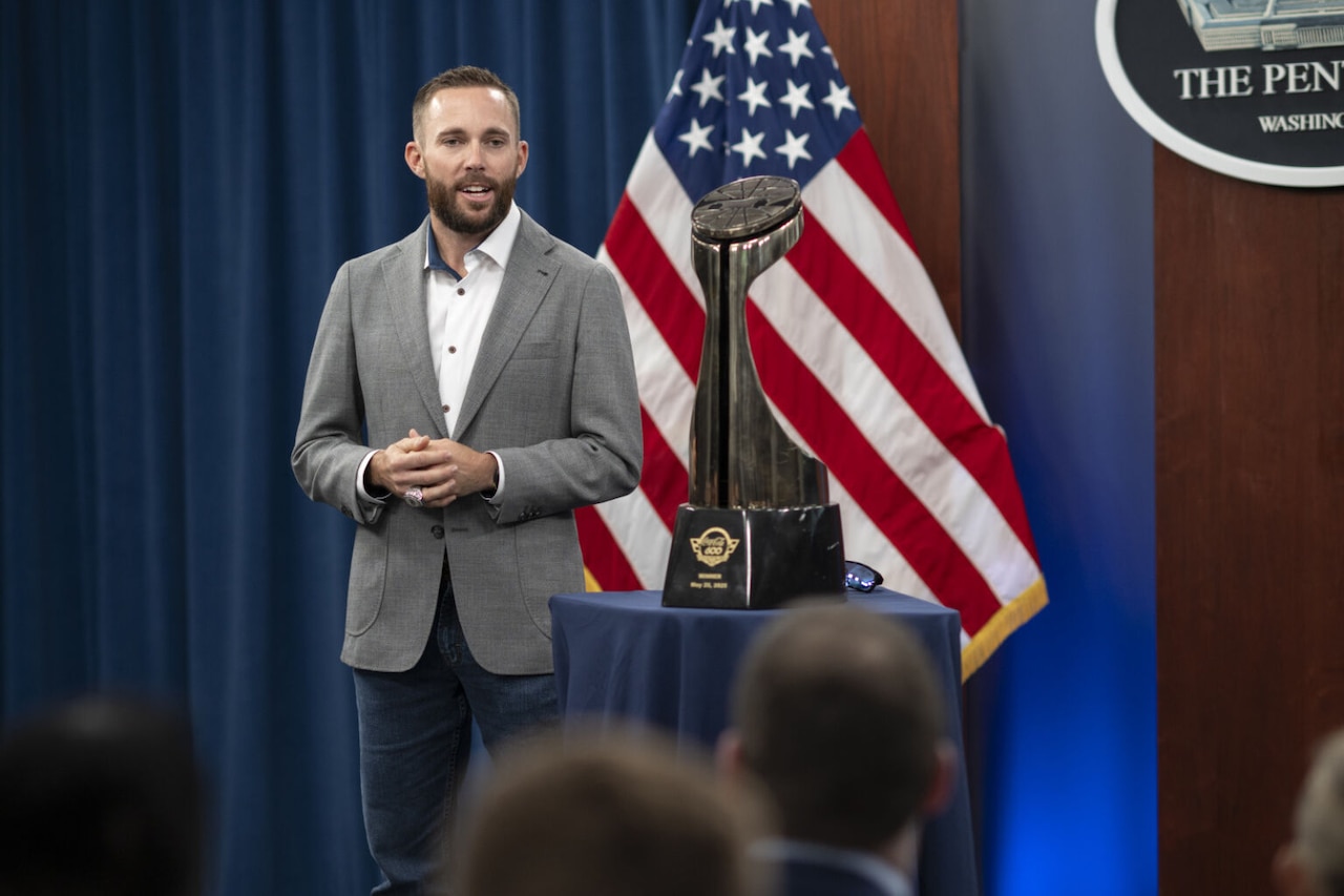 A man wearing business attire stands next to a trophy and speaks to a crowd with an American flag in the background.