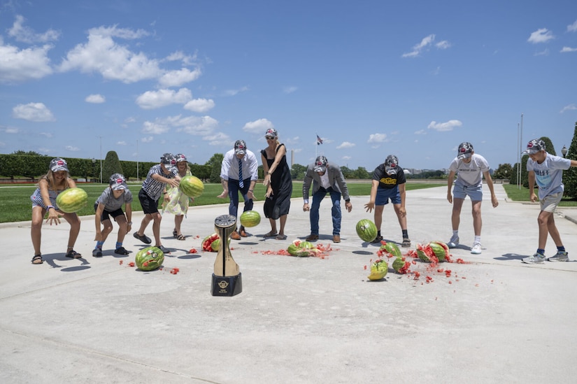 A group of children and adults wearing Coca-Cola 600 hats drop watermelons in unison outside the Pentagon, with the Coca-Cola 600 trophy prominently placed on the ground in front of them.