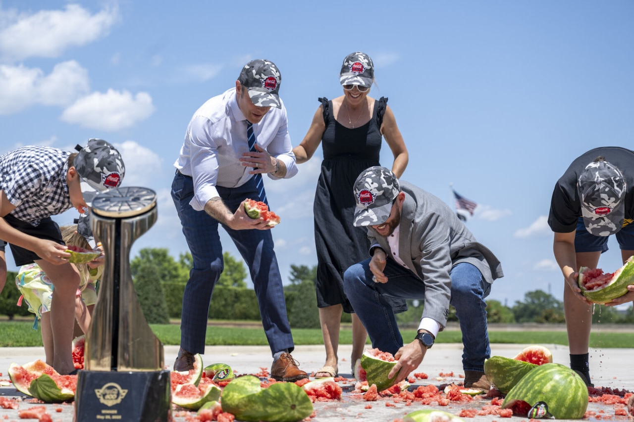 Adults and children crouch on the ground, picking up pieces of smashed watermelon while wearing Coca-Cola 600 hats, with the trophy in the foreground.