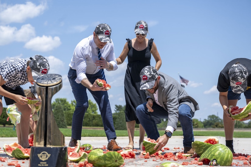 Adults and children crouch on the ground, picking up pieces of smashed watermelon while wearing Coca-Cola 600 hats, with the trophy in the foreground.