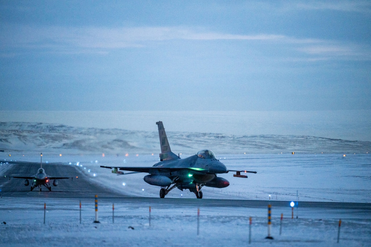Two military aircraft move down a runway, which is surrounded by snow.