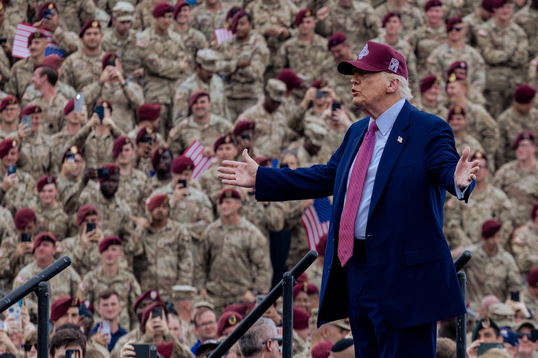 President Donald J. Trump gestures while standing on an outdoor stage speaking to a seated group of soldiers.