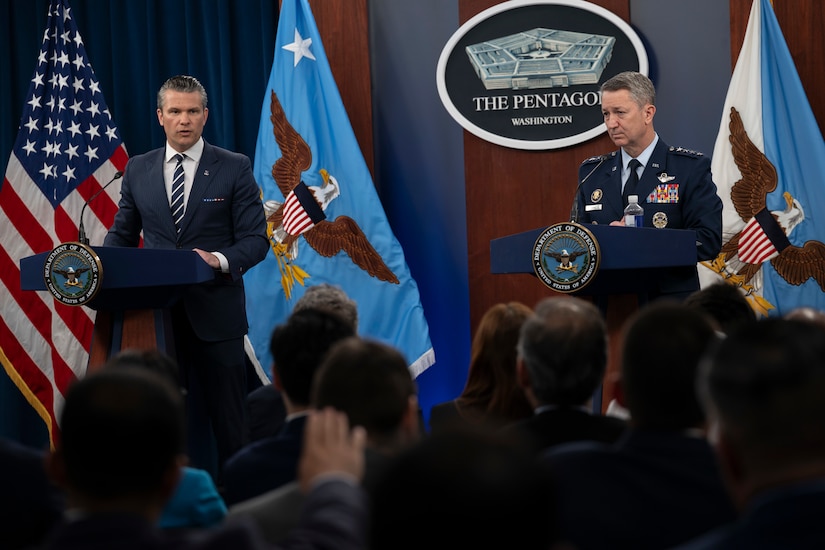 Two men, one in a business suit and the other in a dress military uniform, are standing across from each other behind podiums, speaking to a room full of people sitting down with their backs to the camera.