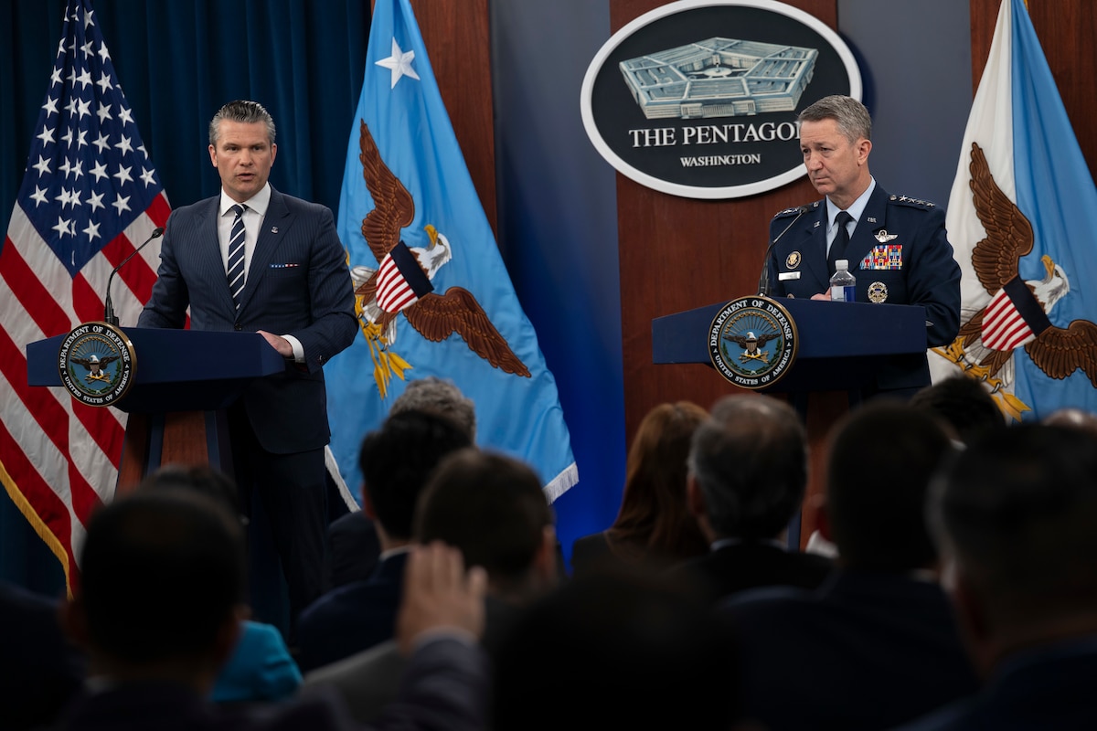 Two men, one in a business suit and the other in a dress military uniform, are standing across from each other behind podiums, speaking to a room full of people sitting down with their backs to the camera.