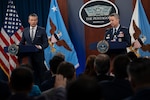 Two men, one in a business suit and the other in a dress military uniform, are standing across from each other behind podiums, speaking to a room full of people sitting down with their backs to the camera.