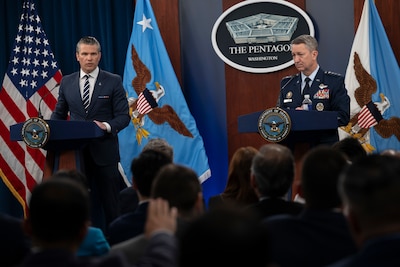 Two men, one in a business suit and the other in a dress military uniform, are standing across from each other behind podiums, speaking to a room full of people sitting down with their backs to the camera.