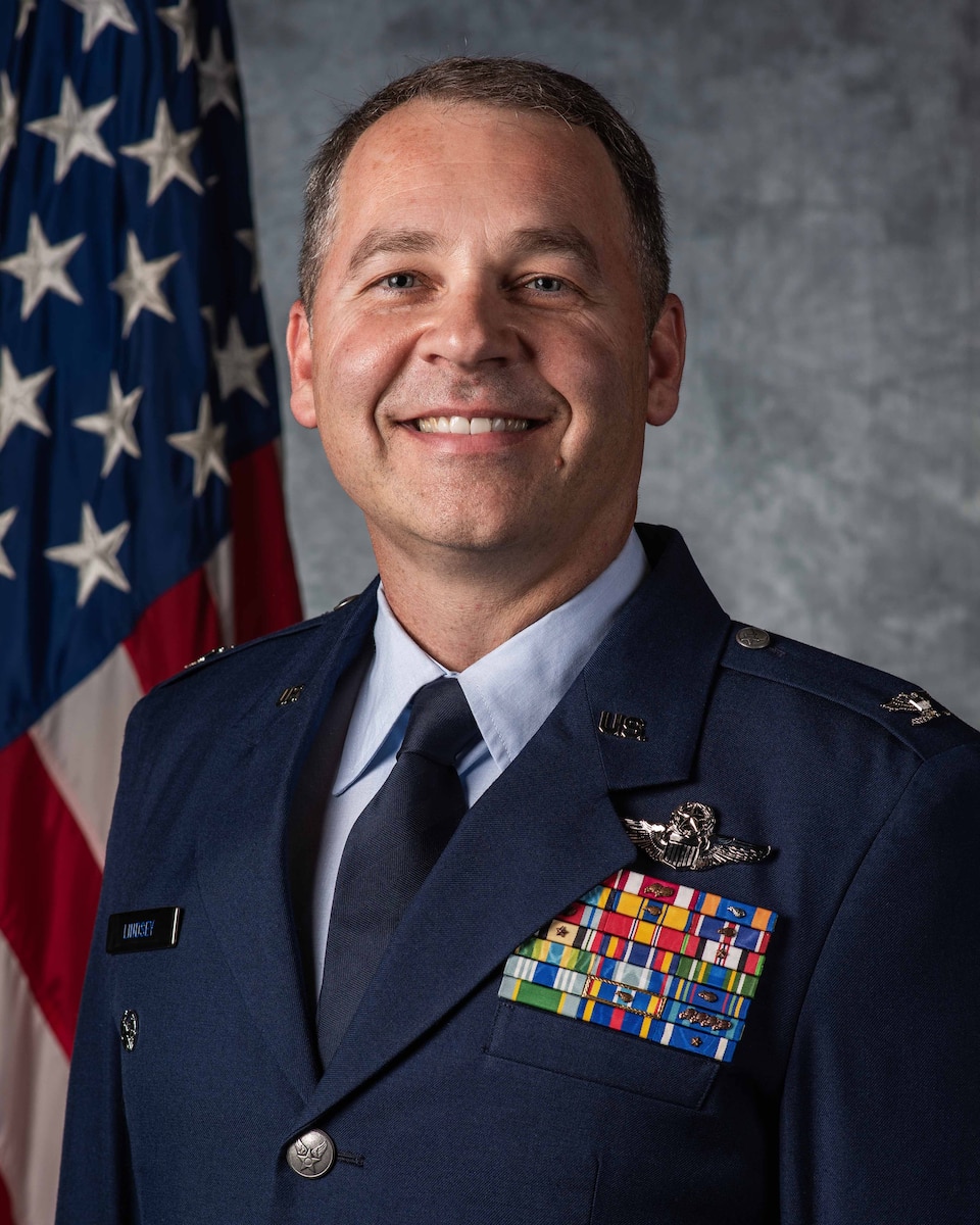 A male Air Force colonel wearing a blues jacket with pilot wings stands and smiles directly into the fame as he stands before an American flag and a cloudy grey background.