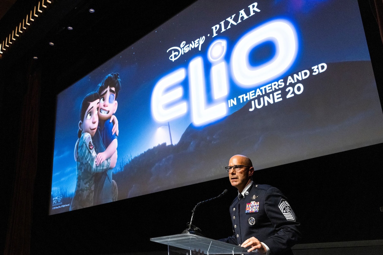 A man in a military dress uniform stands behind a lectern. Behind him is a large movie screen with the words "Disney Pixar, Elio, In Theaters and 3D June 20."