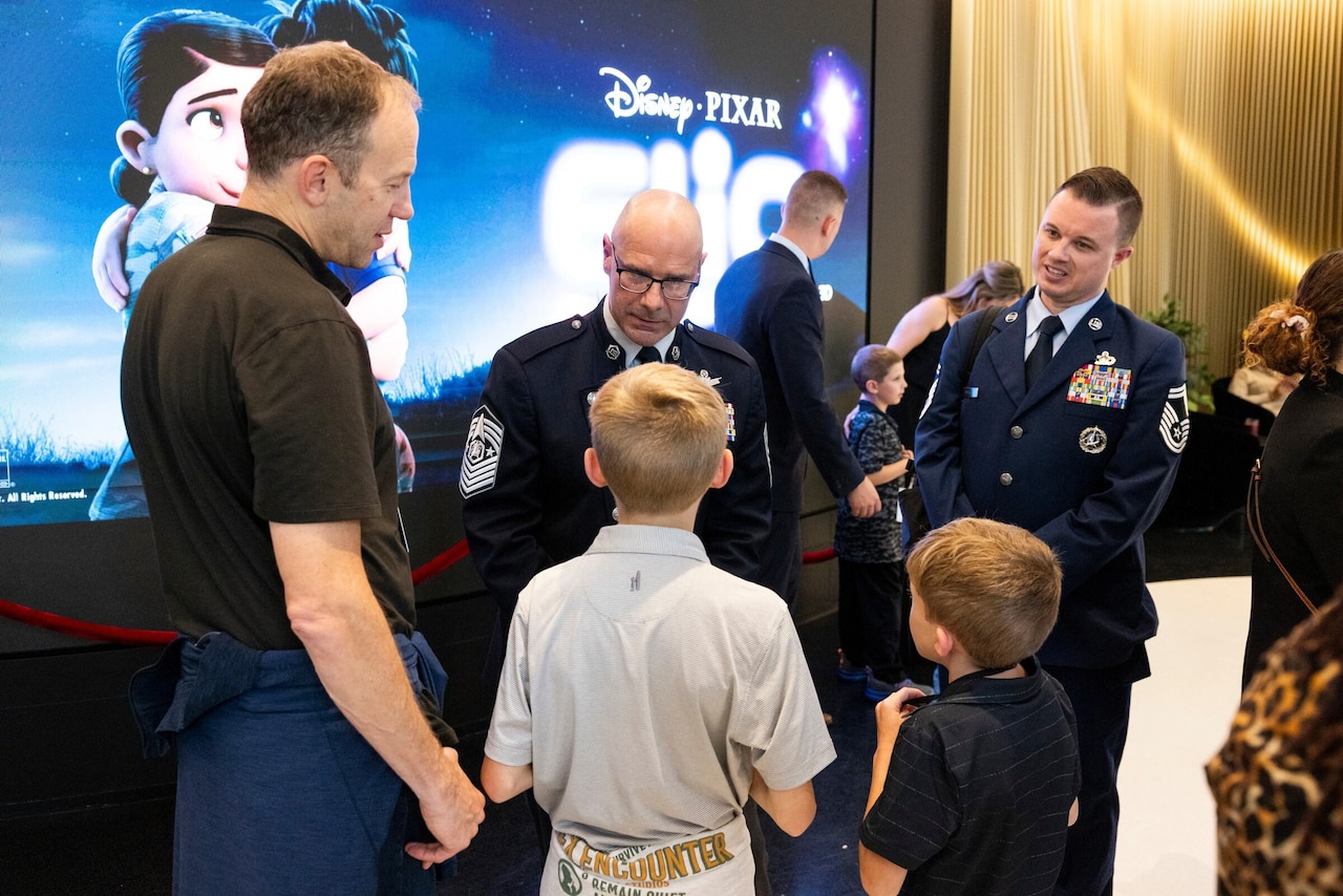 A man in a military dress uniform talks with two children in front of him, while a man in civilian clothes stands beside the children. Another man in a military dress uniform is standing to the side. Behind them is a large movie screen with the words "Disney Pixar, Elio.”