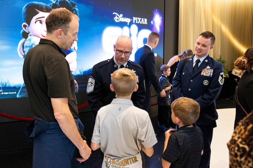 A man in a military dress uniform talks with two children in front of him, while a man in civilian clothes stands beside the children. Another man in a military dress uniform is standing to the side. Behind them is a large movie screen with the words "Disney Pixar, Elio.”