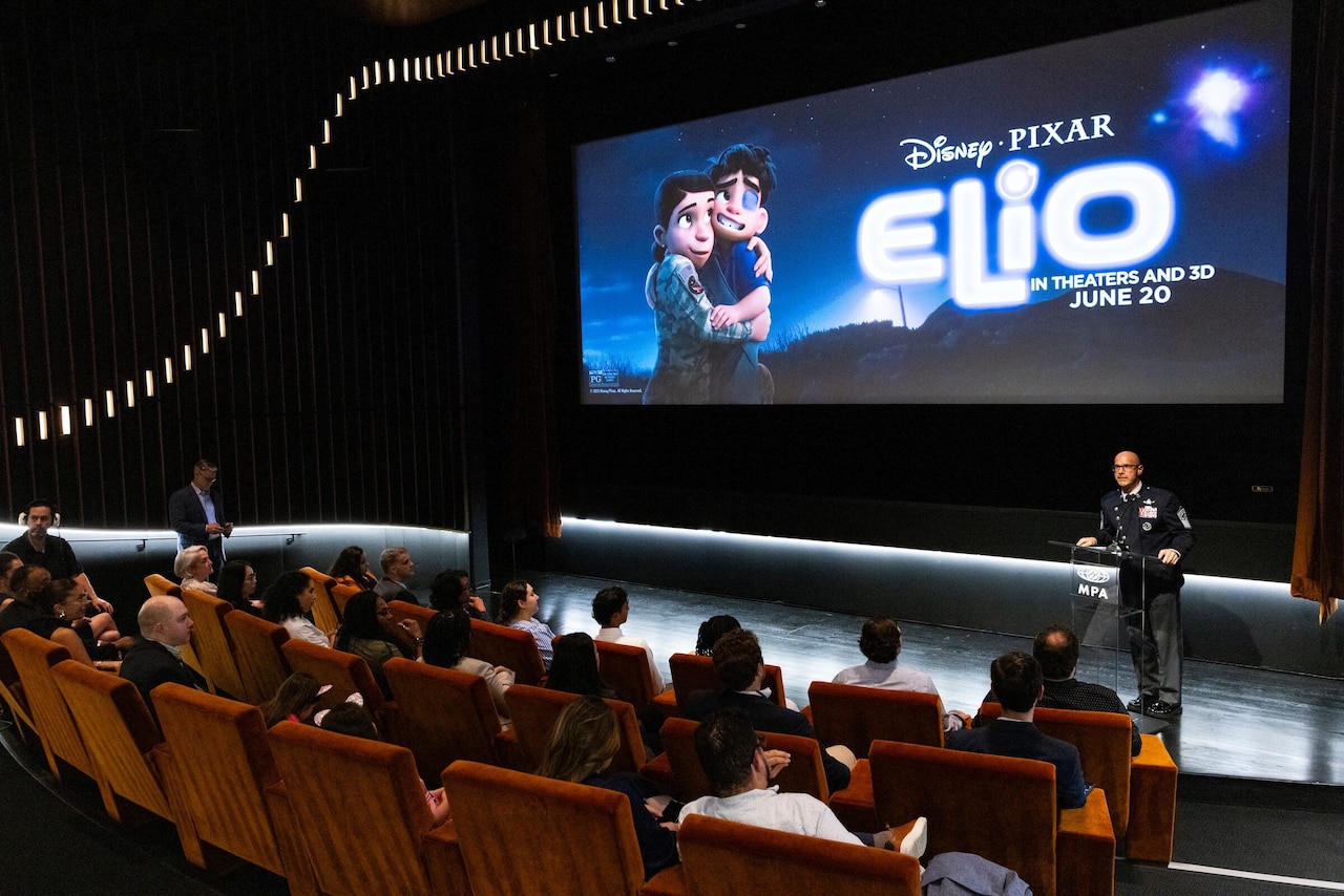 A man in a military dress uniform stands behind a lectern as several people in cvilian attire sit in three rows of movie seats in front of him. Behind him is a large movie screen with the words "Disney Pixar, Elio, In Theaters and 3D June 20."