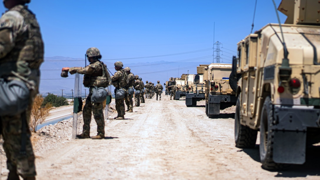 Soldiers holding shields line up on a dirt road with military vehicles parked on the road and mountains in the background.