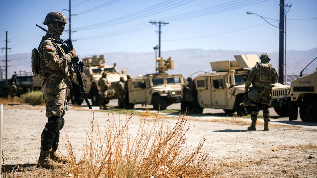 Soldiers in combat gear stand near military vehicles in a desert-like area with power lines and mountains in the background.