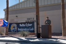 An Airman at a podium giving a speech in front of a military facility.