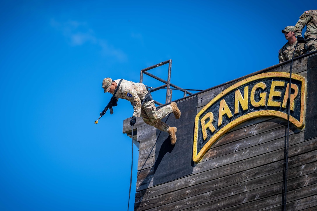 A soldier attached to a rope and holding a rifle walks down a wooden wall outdoors as two other soldiers stand on top of the wall.