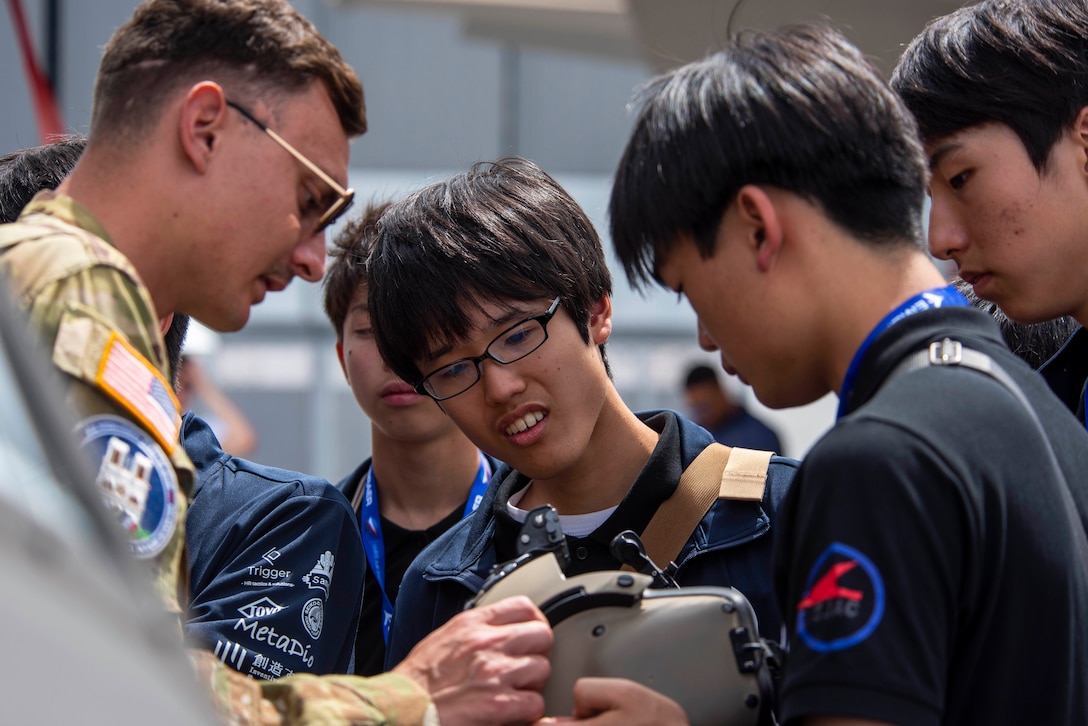 A soldier shows a group of students how a flight helmet works as one student holds the helmet.