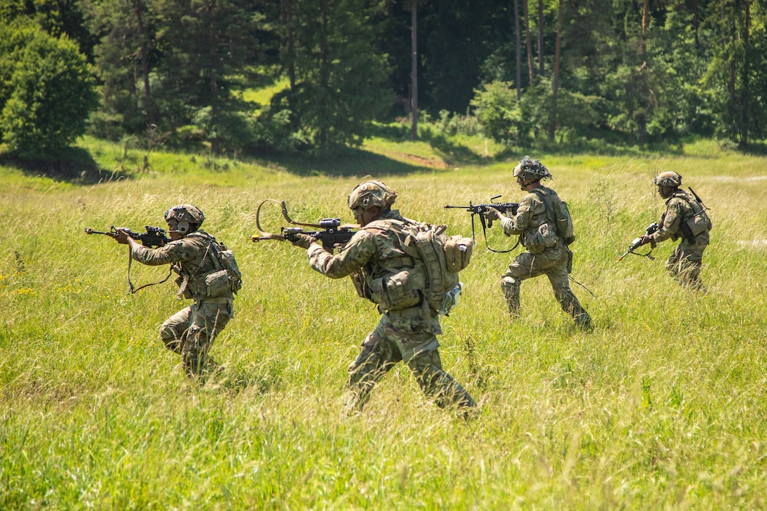 Four soldiers in combat gear move across a grassy field with trees in the background on a sunny day.