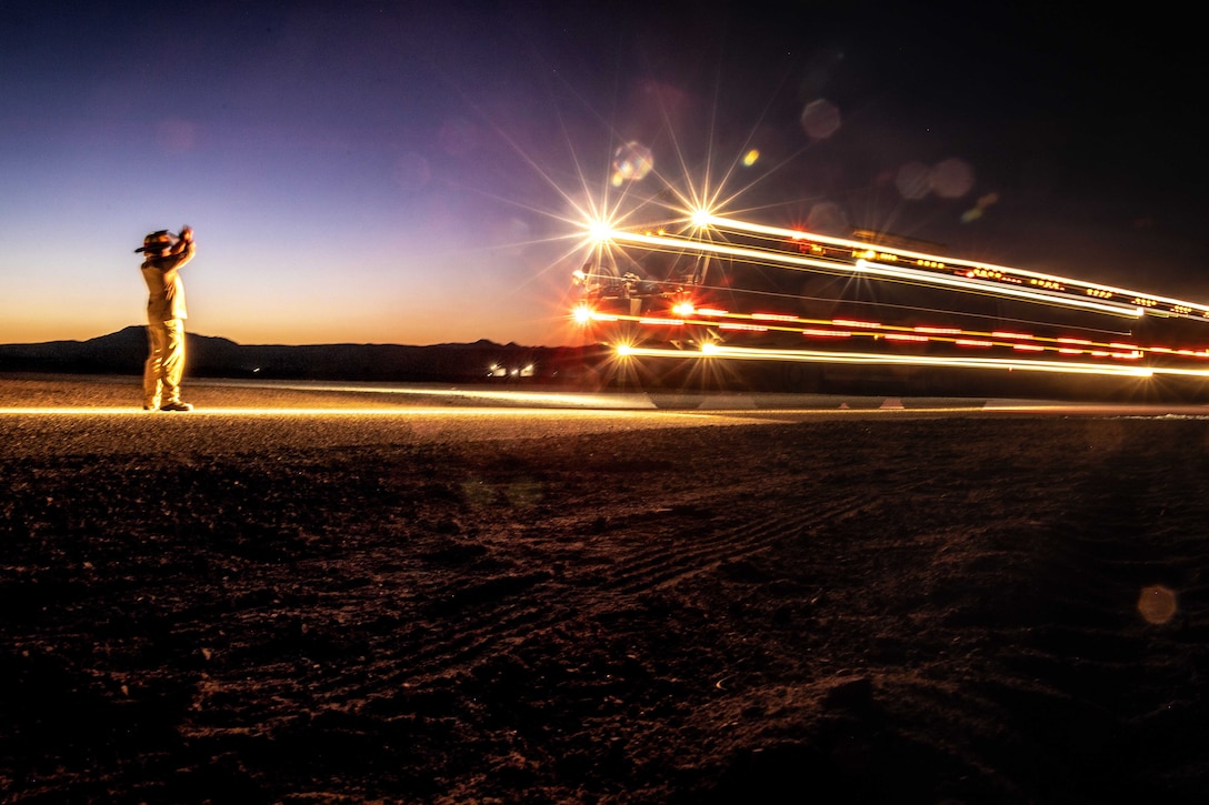 A Marine directs a rescue and firefighting vehicle with its lights on, which are creating a streaming effect with a dark sky in the background.