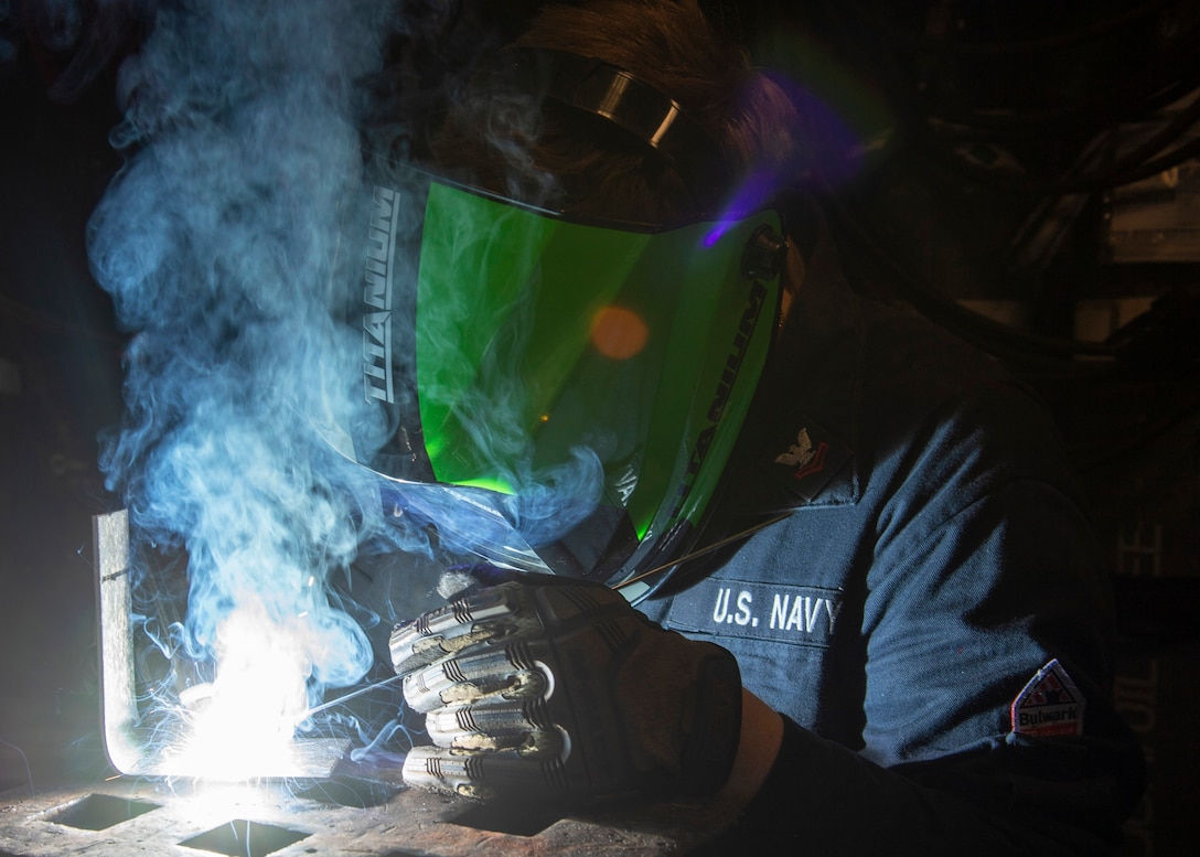 A sailor wearing protective head and hand gear welds an "L"-shaped piece of metal.