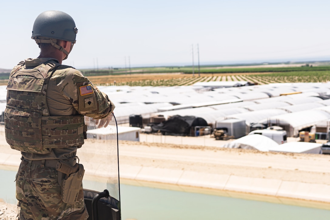 A soldier holding a shield looks out over a tent complex with vehicles and equipment; a field is in the background.