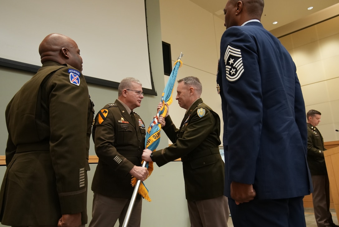 four men in uniform stand on stage, with one passing a flag to another in the center