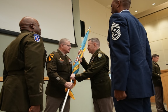 four men in uniform stand on stage, with one passing a flag to another in the center