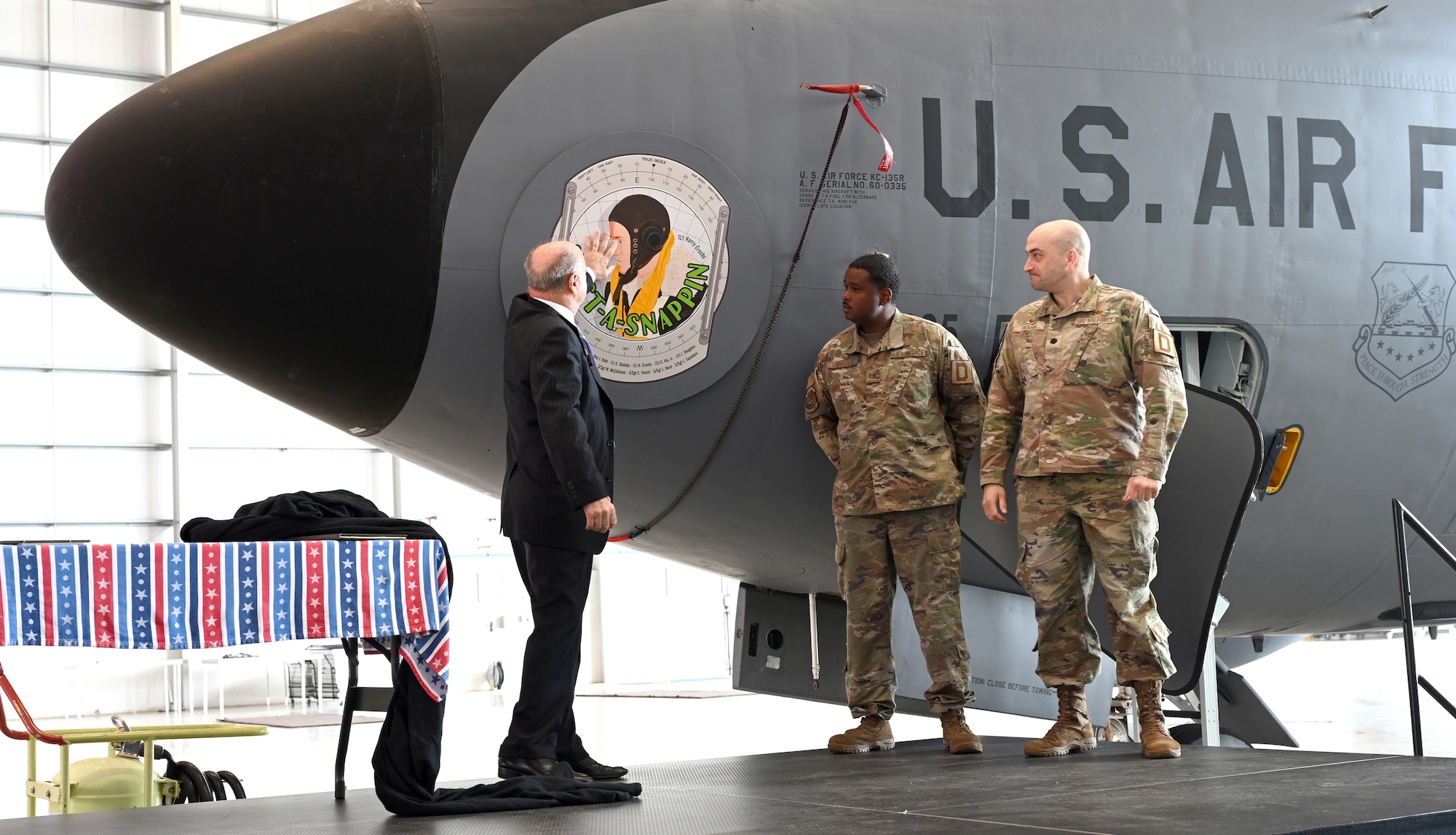 Steve Crosby, left, son of then-1st Lt. Harry Crosby, World War II and 100th Bomb Group navigator, touches the latest legacy nose art “Just-A-Snappin,” belonging to the 100th Air Refueling Wing, as Lt. Col. Scott Engman, right, 100th Logistics Readiness Squadron commander, and Tech. Sgt. Devan Conerly, 100th Aircraft Maintenance Squadron Airframe, Powerplant, General NCO-in-charge, witness the nose art unveiling during a ceremony at RAF Mildenhall, England, June 9, 2025. The nose art, designed by Gary Rogers, 100th ARW Public Affairs graphic designer, focuses on the profile of Steve’s father and incorporates a background of a navigator’s dead-reckoning computer. It also bears the names of the crew who flew the Bremen mission over Germany in October 1943. (U.S. Air Force photo by Karen Abeyasekere)