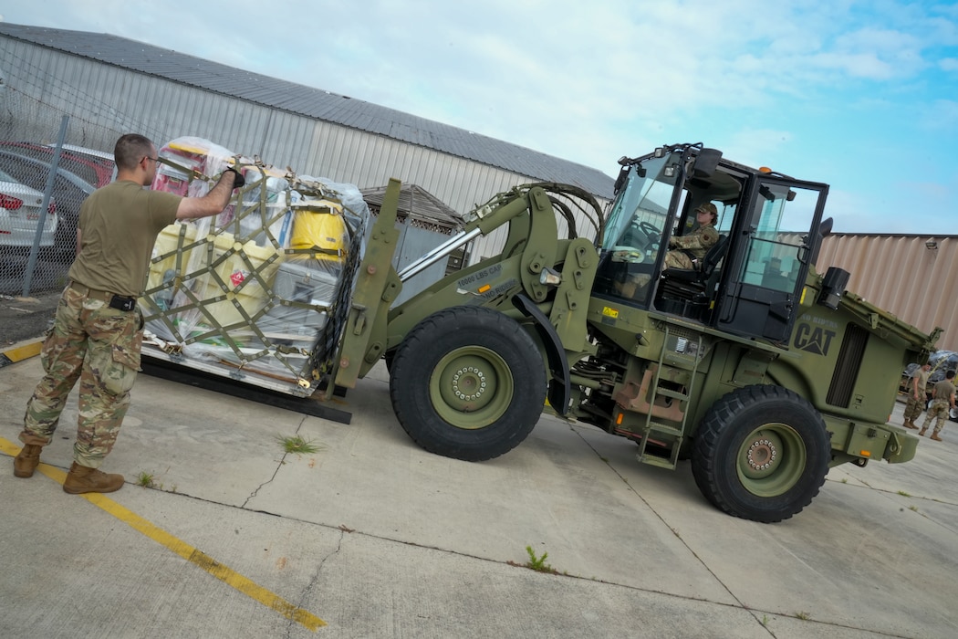 Photo shows large equipment carrying pallet being driven by woman in uniform while being directed by man in uniform